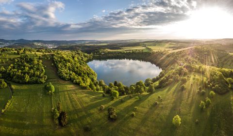 Ein malerischer See umgeben von üppigen Wiesen und Bäumen. Die Sonne geht über der Landschaft auf und wirft sanfte Strahlen auf das Wasser.
