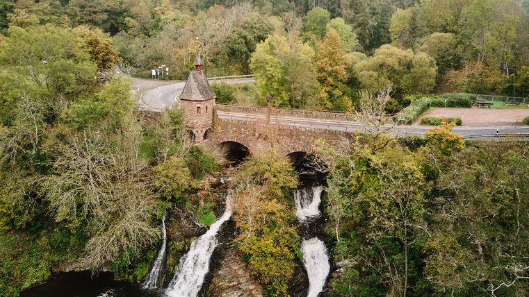 Der Elzbach-Wasserfall fließt unter einer alten Steinbrücke mit einem kleinen Turm hindurch. Die Brücke führt durch einen Wald. Die Bäume haben grüne und gelbe Blätter. Das Wasser fällt in mehreren Stufen nach unten.