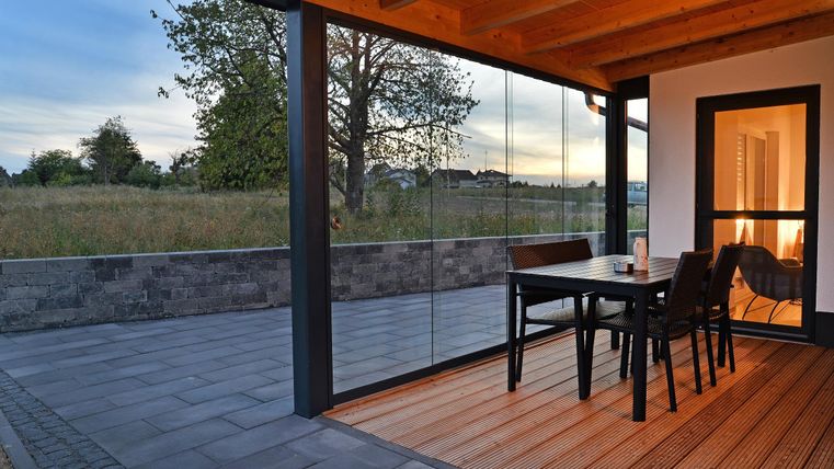 Ein modern gestalteter Außenbereich mit einem Tisch und Stühlen auf einer Terrasse. Der Blick geht auf eine grüne Wiese und einen bewölkten Himmel.