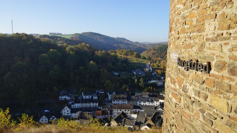 Ein Blick auf eine malerische Stadt im Tal, umgeben von sanften Hügeln. Im Vordergrund ist ein Teil einer alten Mauer zu sehen.