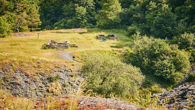 Blick von oben auf eine grüne Lichtung mit Ruinen aus Bruchstein, umgeben von dichtem Wald und Schieferhängen.