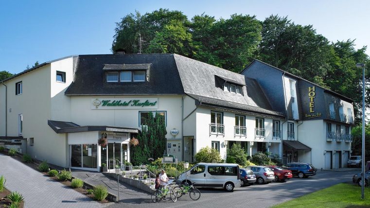 Ein modernes Hotelgebäude umgeben von Bäumen. Auf dem Parkplatz sind Autos und Fahrräder zu sehen.