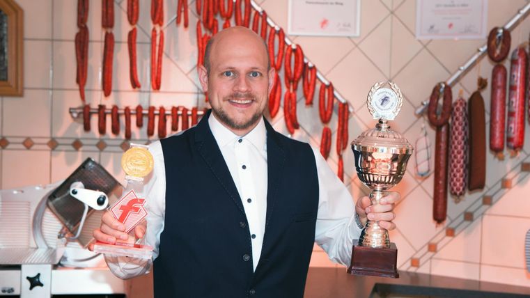 Ein stolzer Mann in einem Restaurant hält einen Pokal und eine Auszeichnung in der Hand. Im Hintergrund hängen verschiedene Wurstsorten an der Wand.