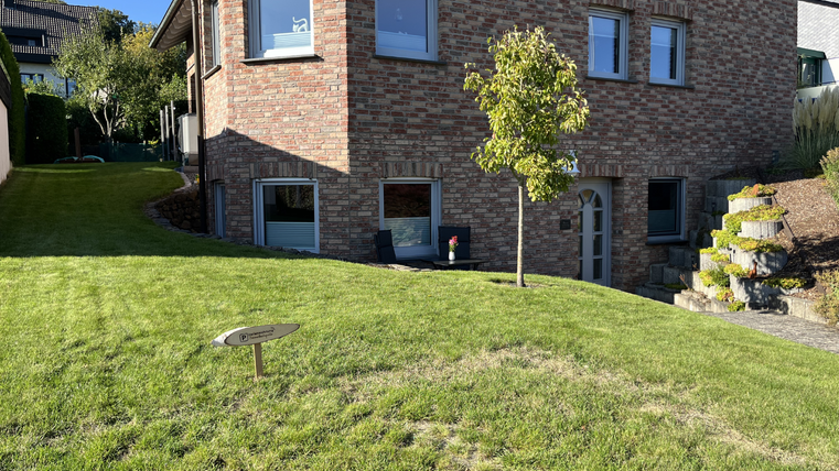 Ein Haus mit Ziegelfassade und großen Fenstern steht in einem schönen Garten. Der Rasen ist gepflegt und es gibt einen kleinen Baum sowie eine Sitzgelegenheit neben dem Eingang.