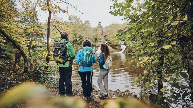Drei Wanderer stehen am Ufer eines ruhigen Flusses und betrachten die Landschaft. Um sie herum ist eine herbstliche Szenerie mit bunten Bäumen.