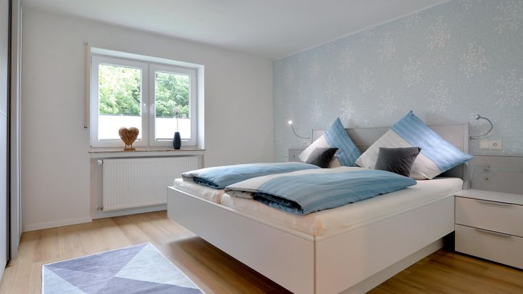 Ein modernes Schlafzimmer mit einem großen Doppelbett und hellen Farben. Der Raum hat große Fenster und einen gemütlichen Teppich.