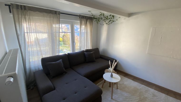 Ein modernes Wohnzimmer mit einem bequemen dunkelgrauen Sofa und einem kleinen weißen Tisch. Große Fenster lassen viel Tageslicht herein und die Wände sind hell gestrichen.