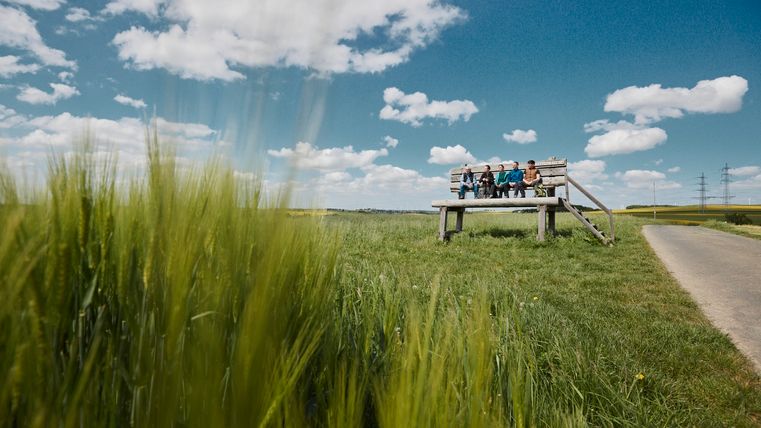 Mehrere Personen sitzen auf einer übergroßen Holzbank an einem Feldweg inmitten einer weiten, grünen Landschaft unter einem blauen Himmel mit weißen Wolken. Im Vordergrund wiegen sich Ähren im Wind.