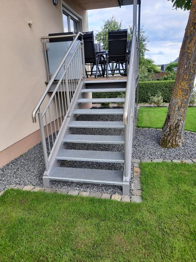 Eine moderne Treppe aus Metall führt zu einem Außenbereich mit Möbeln. Der Garten ist gepflegt und grün, begleitet von Kies.