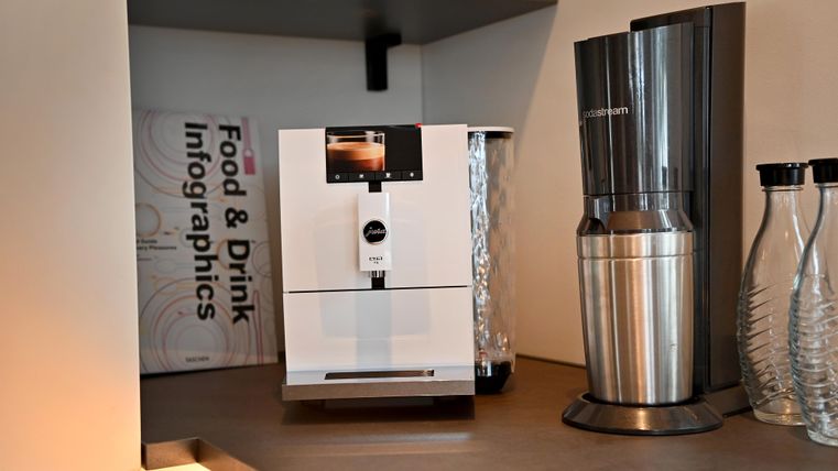 Eine moderne Kaffeemaschine steht auf einer Arbeitsplatte in einer stilvollen Küche. Im Hintergrund sind Flaschen und ein Buch über Essen und Getränke sichtbar.