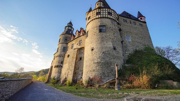 Eine beeindruckende Burg mit hohen Türmen und historischen Elementen. Umgeben von grüner Landschaft unter einem klaren Himmel.