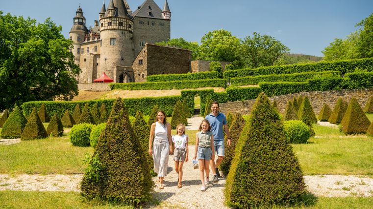 Eine Familie steht in einem Garten mit kunstvoll geschnittenen Hecken. Im Hintergrund ist ein beeindruckendes Schloss zu sehen.
