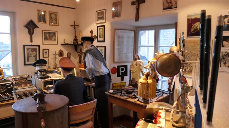 Een historische ruimte met verschillende kunstvoorwerpen en portretten aan de muren. Twee personen interageren te midden van een verzameling antiquiteiten en souvenirs.