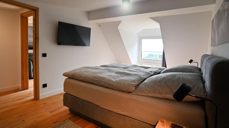 Ein modernes Schlafzimmer mit einem großen Bett und einem Fernseher an der Wand. Die Wände sind hell und das Fenster bietet einen Blick nach draußen.