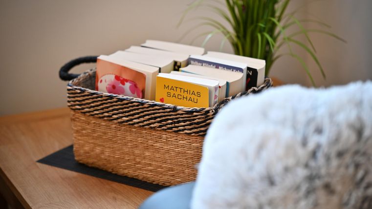 A basket with several books is on a table. In the background, a plant is visible.