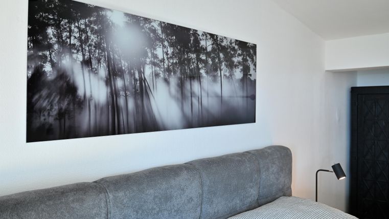 Ein modernes Schlafzimmer mit einem grauen Bett und einer Kunstfotografie von einem Wald an der Wand. Die Atmosphäre wirkt ruhig und einladend.