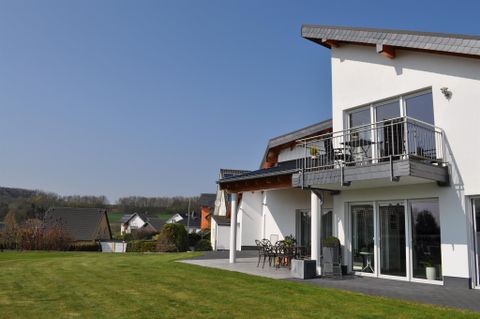 Ein modernes Haus mit einem schönen Balkon und großen Fenstern. Der Garten ist gepflegt und grün, umgeben von sanften Hügeln.