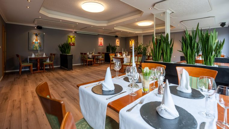 Ein elegantes Restaurant mit stilvoll gedeckten Tischen. Die Innenausstattung ist modern und einladend, umgeben von Pflanzen und Kunstwerken.