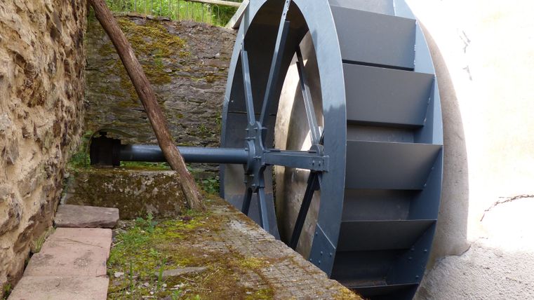 Une roue hydraulique se trouve contre un vieux mur. Elle est bleu foncé et fait partie d'un système hydrolique historique.