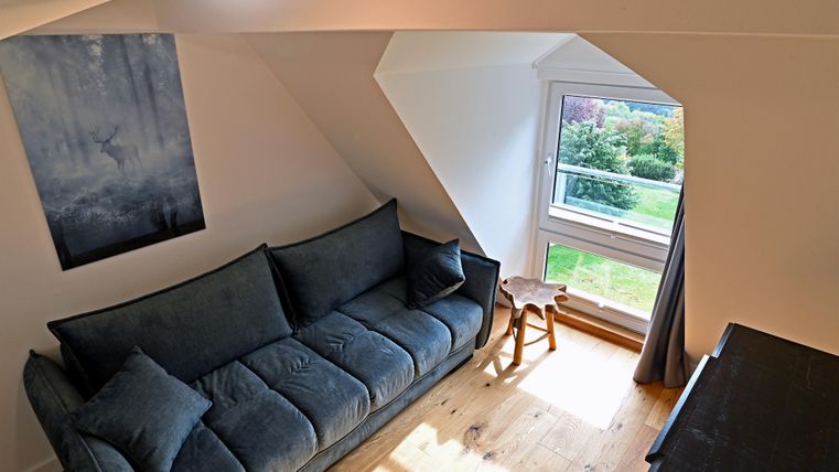 Ein modernes Wohnzimmer mit einem blauen Sofa und einem kleinen Tisch. Durch das Fenster sieht man ins Grüne.