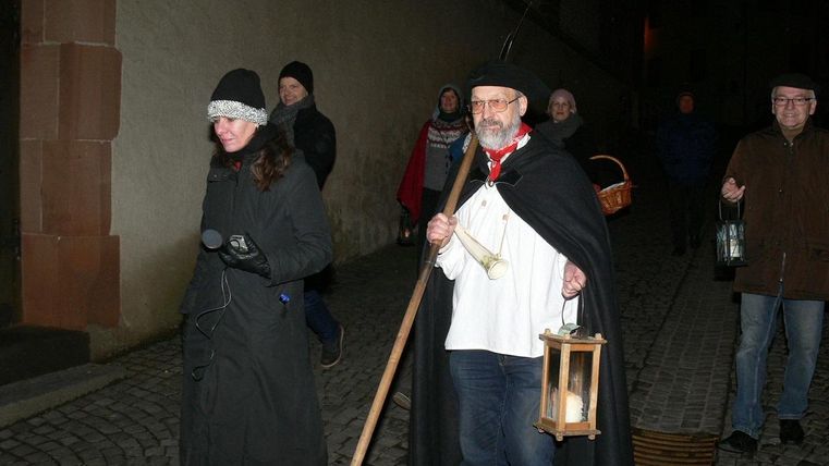 Eine Gruppe von Menschen geht nachts eine gepflasterte Straße entlang. Der Mann in der Mitte trägt eine Laterne und einen langen Umhang.