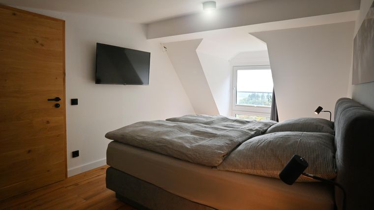 Ein modernes Schlafzimmer mit einem großen Bett und einem Fernseher an der Wand. Das Zimmer hat ein freundliches Ambiente mit einem Fenster, das Tageslicht hereinlässt.