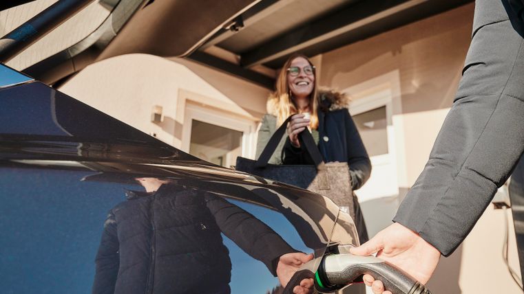 Eine lächelnde Frau steht neben einem E-Auto und beobachtet das Aufladen. Der Fokus liegt auf dem Ladekabel, das mit dem Auto verbunden ist.