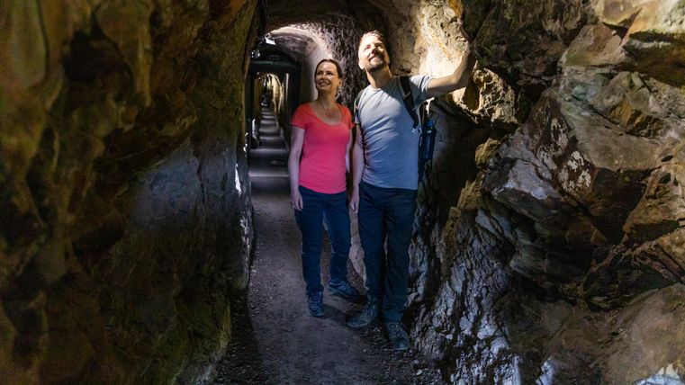 Ein Paar steht in einem schmalen, steinernen Tunnel und schaut begeistert umher. Die Wände des Tunnels sind rau und natürlich, während das Licht eine geheimnisvolle Atmosphäre schafft.