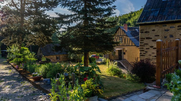 Un jardin pittoresque avec des plantes et des arbres colorés. En arrière-plan, des bâtiments traditionnels sont visibles.