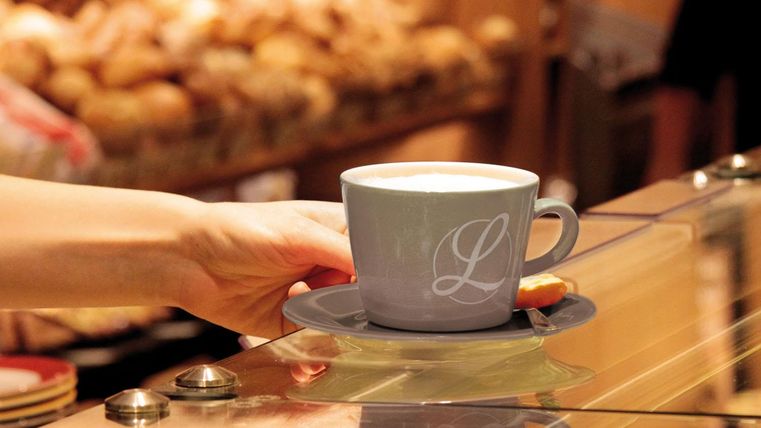 Eine Hand hält eine Tasse Kaffee auf einem Unterteller. Im Hintergrund sind frische Brötchen zu sehen.