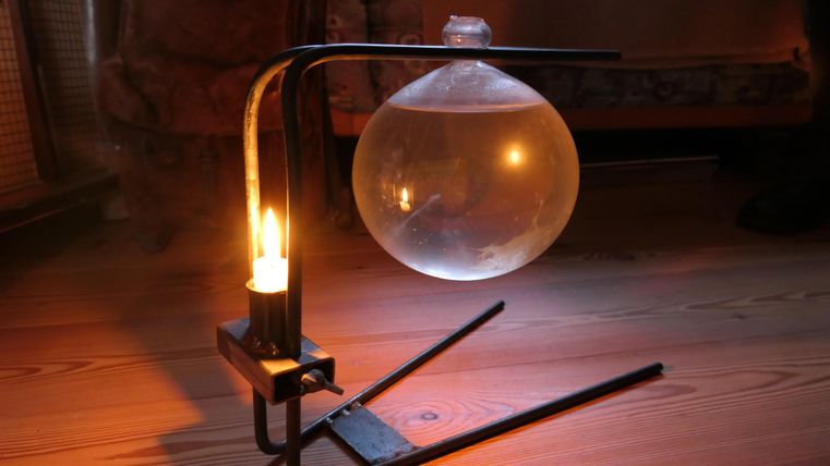 Een elegante lamp met een ronde glazen bol en een brandende kaars. Het licht creëert een warme sfeer in een kamer met een houten vloer.