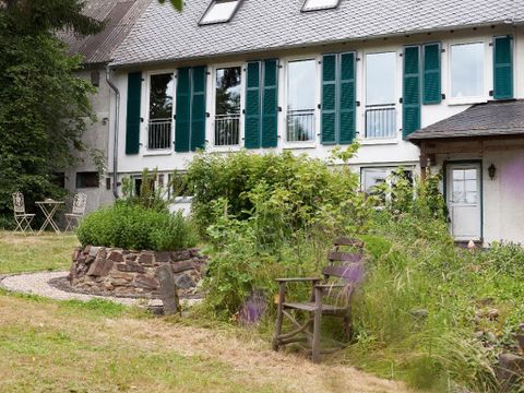 Ein gemütliches Haus mit grünen Fensterläden und einem gepflegten Garten. Im Vordergrund steht eine Holzbank umgeben von Pflanzen.