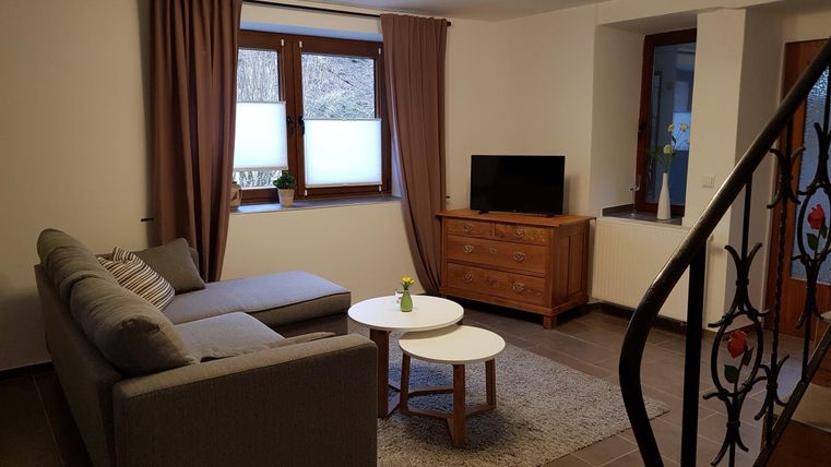 Ein gemütliches Wohnzimmer mit einem grauen Sofa, einem weißen Couchtisch und einem Fernseher. Große Fenster bringen natürliches Licht in den Raum.