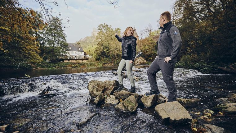 Zwei Personen stehen an einem kleinen Wasserfall und genießen die Natur. Im Hintergrund sind Bäume und ein Haus zu sehen.