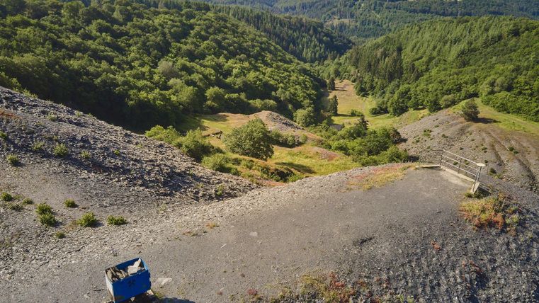 Eine malerische Landschaft mit sanften Hügeln und üppigem Grün. Im Vordergrund ist ein blauer Wagen auf einem kiesigen Weg zu sehen.