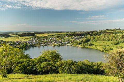 Eine malerische Landschaft mit einem klaren See und sanften Hügeln. Im Hintergrund sieht man ein kleines Dorf umgeben von Bäumen.