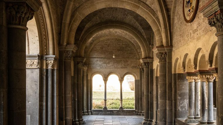 Ein historischer Innenhof mit hohen Bögen und Säulen. Das Licht fällt sanft durch die Fenster und beleuchtet die Steinmauern.