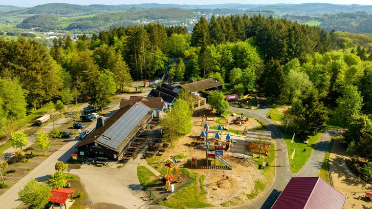 Ein luftiger Blick auf einen Spielplatz umgeben von Bäumen und Hütten. Die freundliche Umgebung lädt Familien zu Erholung und Spiel ein.