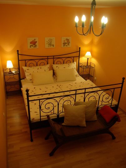 Ein gemütliches Schlafzimmer mit einem Eisengestellbett und orangefarbenen Wänden. Neben dem Bett stehen zwei Nachttische mit Lampen.