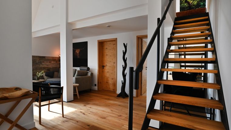 Ein modernes Interieur mit Holztreppe und gemütlichem Sittingbereich. Große Fenster sorgen für viel Tageslicht und eine freundliche Atmosphäre.