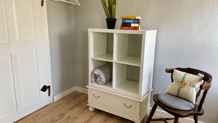 Ein heller Raum mit einem weißen Regal und einem Holzstuhl. Auf dem Regal stehen einige Bücher und ein Kaktus.