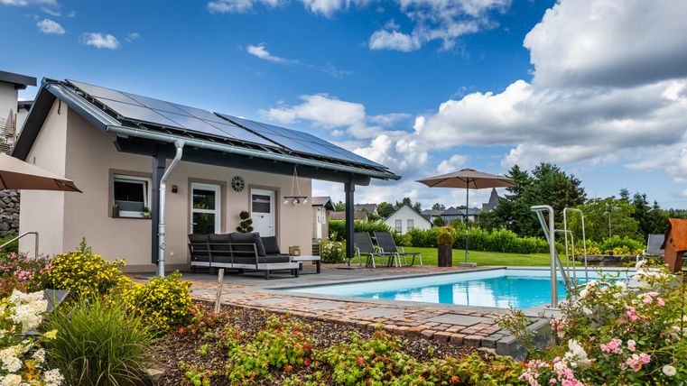 Ein modernes Haus mit Solarpanels und einem Swimmingpool. Der Garten ist schön mit Blumen und einer sonnigen Umgebung gestaltet.