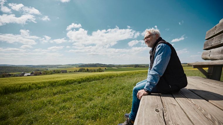 Ein älterer Mann sitzt auf einer Holzbank und schaut in die grüne Landschaft. Der Himmel ist klar mit einigen Wolken.