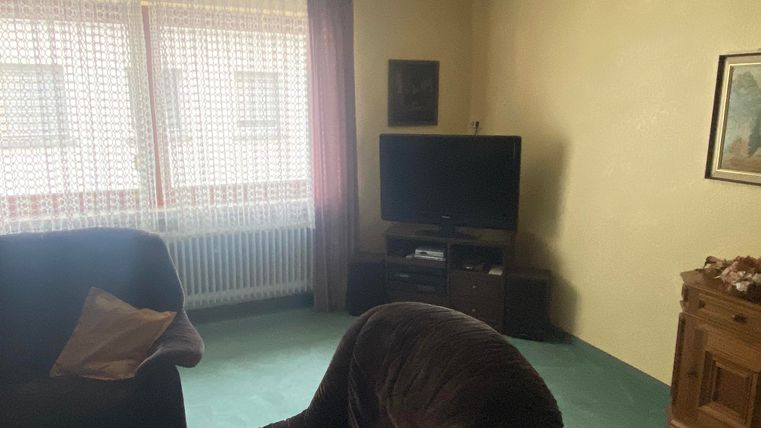 Ein einfaches Wohnzimmer mit einem Fernseher und einem Holzschrank. Es gibt einen bequemen Sessel und große Fenster mit Vorhängen.