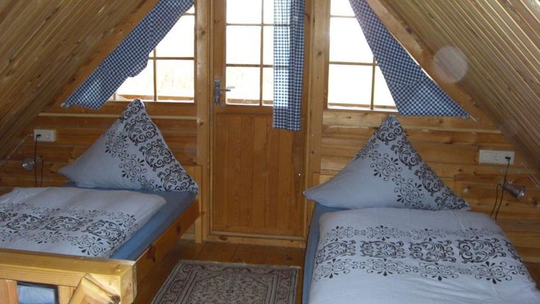 Une chambre mansardée confortable avec deux lits individuels et des coussins bleus. Les murs sont en bois, et une fenêtre laisse entrer beaucoup de lumière du jour.