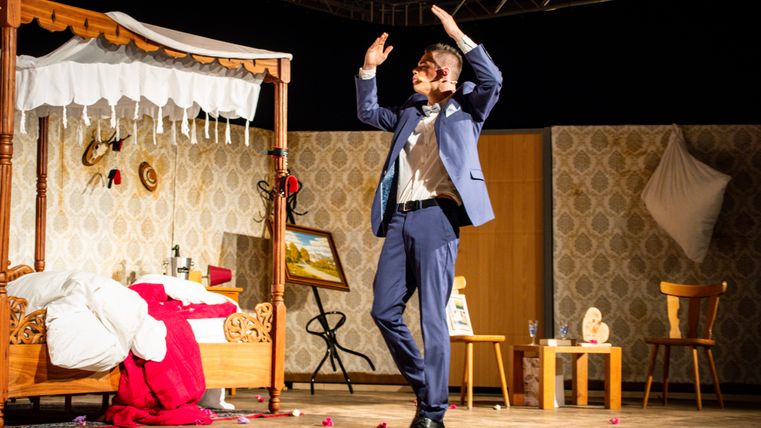 Schauspieler in blauem Anzug auf einer Theaterbühne vor einer Schlafzimmerkulisse während einer Komödienaufführung.
