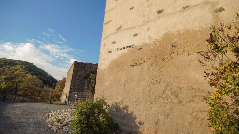 Eine alte Mauer mit sichtbaren Steinen und etwas Bewuchs. Im Hintergrund sind sanfte Hügel und ein blauer Himmel zu sehen.