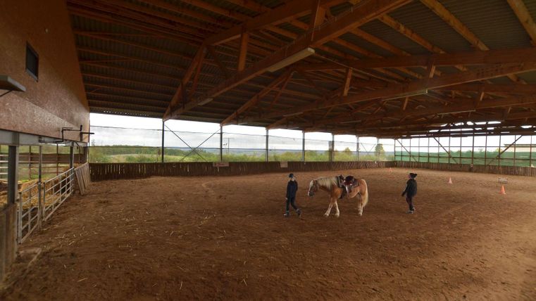 Une carrière équestre avec un grand sol en sable et une structure en bois. Deux personnes s'occupent d'un cheval à l'intérieur.