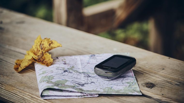 Eine gefaltete Karte liegt auf einem Holztisch, neben einem GPS-Gerät. Ein gelbes Blatt liegt ebenfalls auf dem Tisch.
