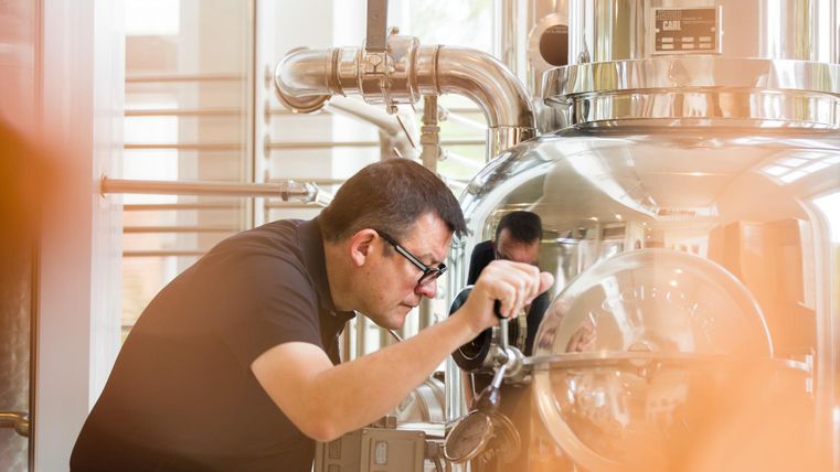 Ein Mann beobachtet aufmerksam einen großen, glänzenden Destillationsbehälter in einer modernen Brennerei. Im Hintergrund sind metallische Rohre und eine helle, offene Umgebung sichtbar.